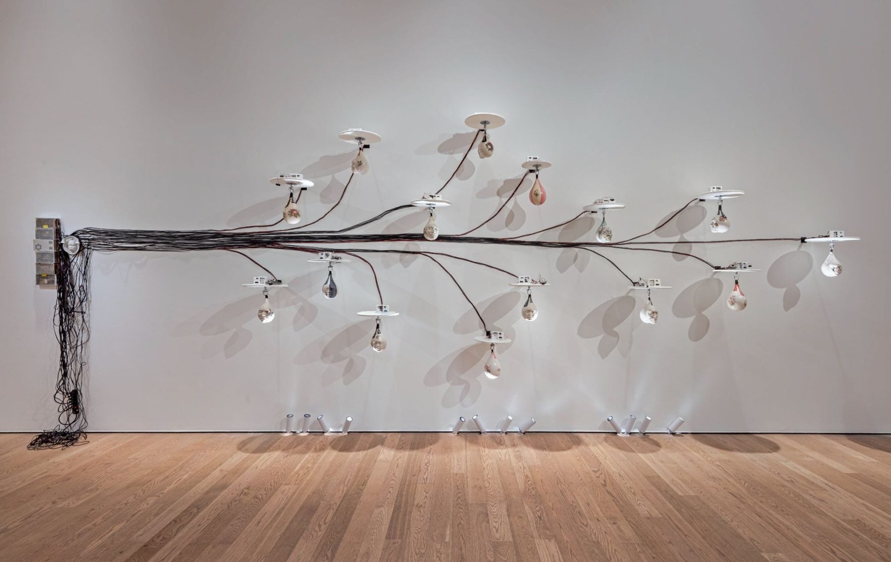 Installation artwork with a long branch-like structure spanning a white wall, featuring multiple suspended light fixtures and dangling components, set against a wooden floor.