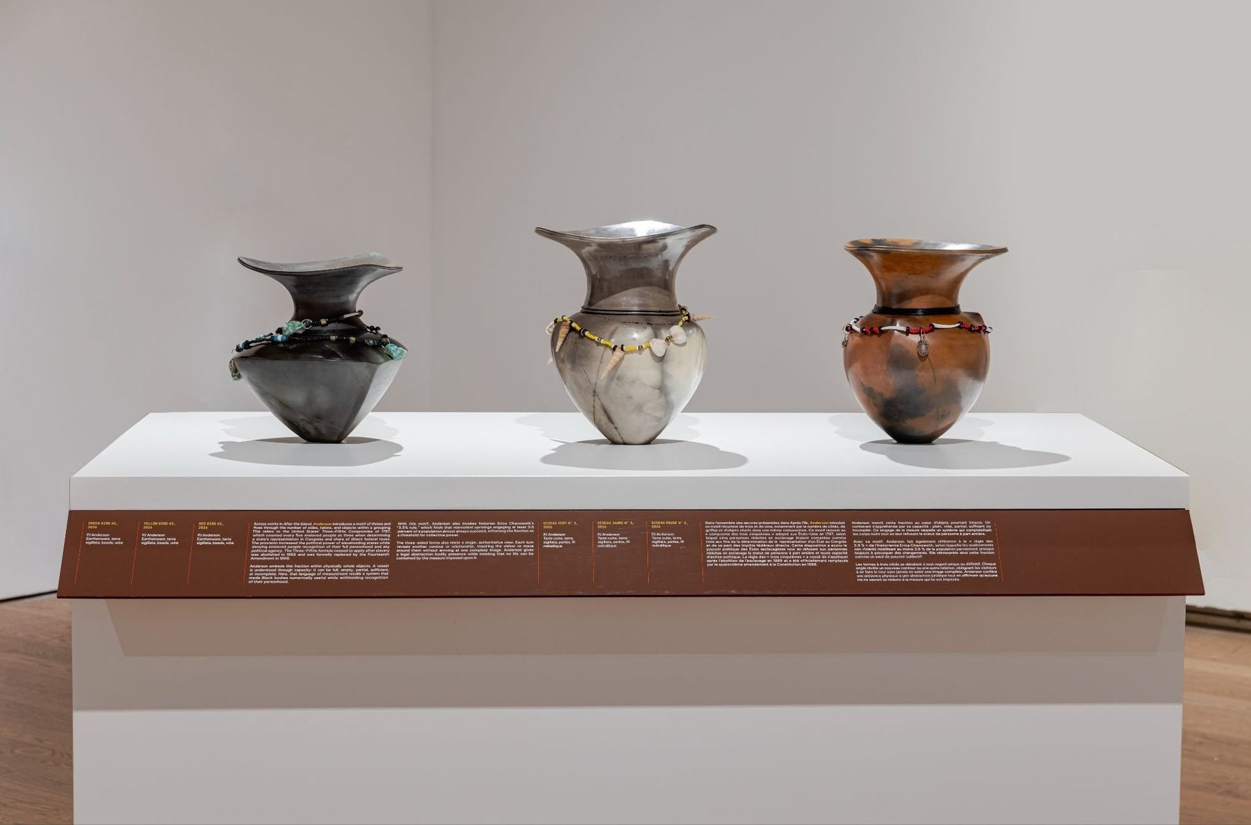 Three ceramic vases on display with beaded necklaces, in a museum pedestal setting.