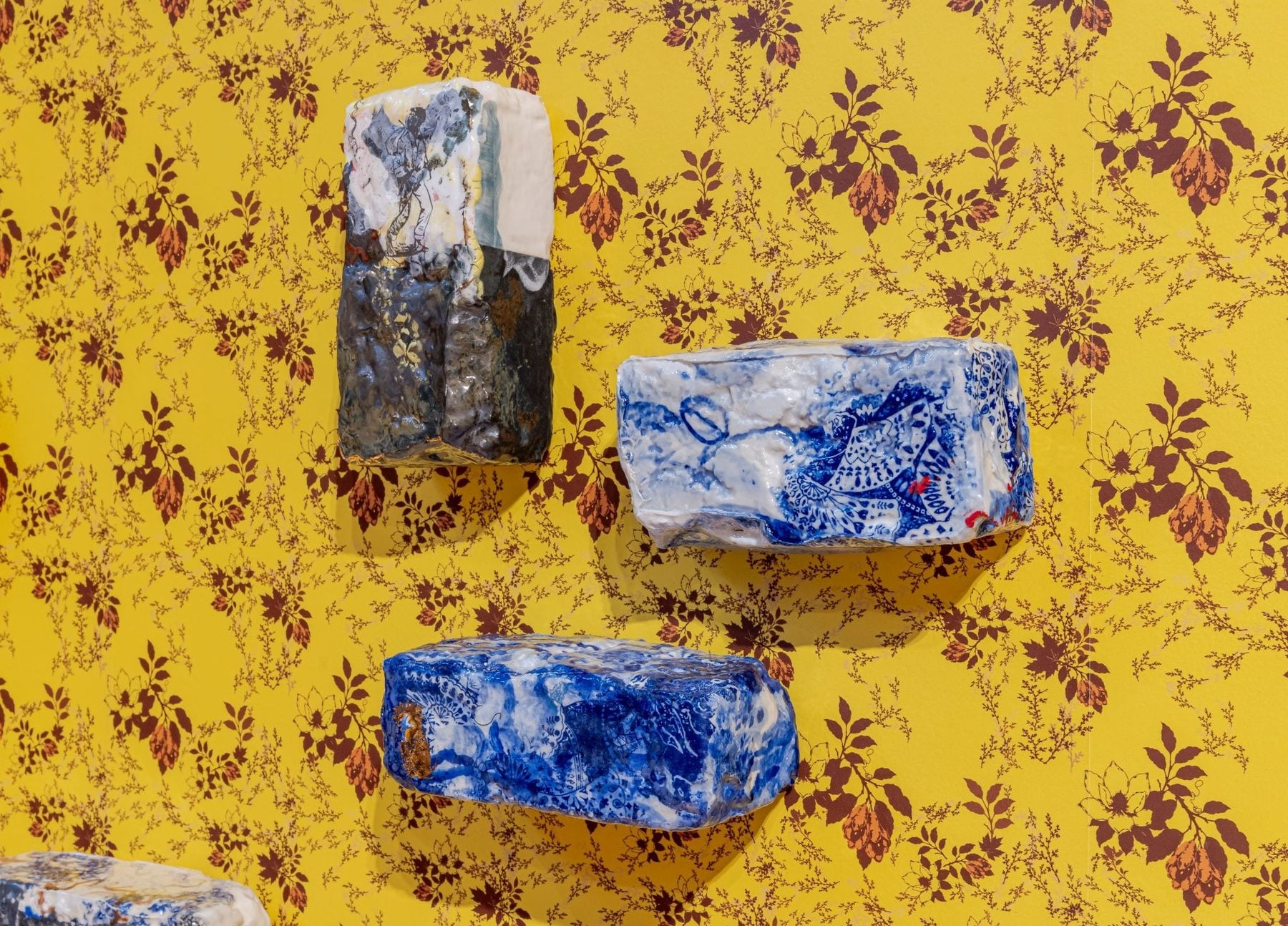 Carved ceramic blocks with blue and white patterns mounted on a yellow floral wallpaper backdrop, part of a Gardiner Museum exhibit exploring Caribbean identity.