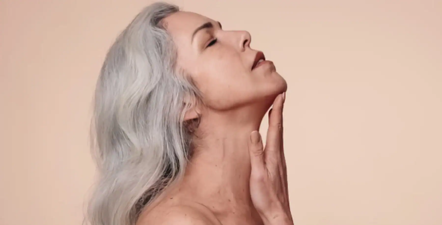 Elegant mature woman with long silver hair gently touching her neck, showcasing natural beauty and confidence in a neutral background.
