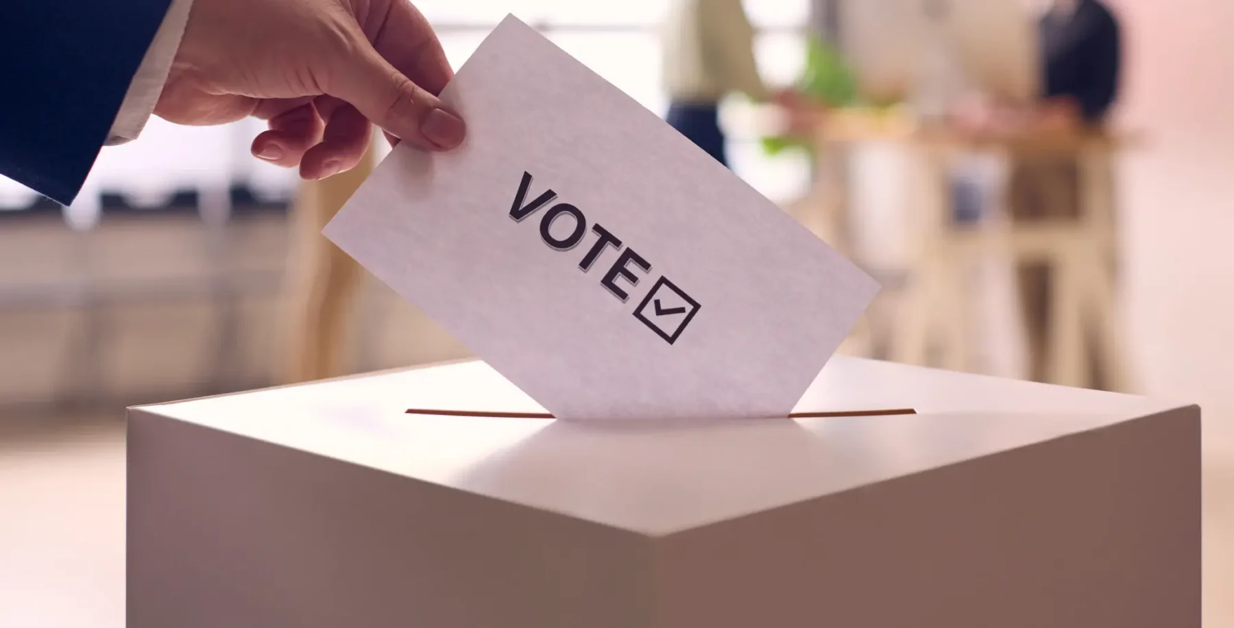 Voter casts ballot into a mailbox slot during an election, with focus on the vote envelope labeled VOTE.