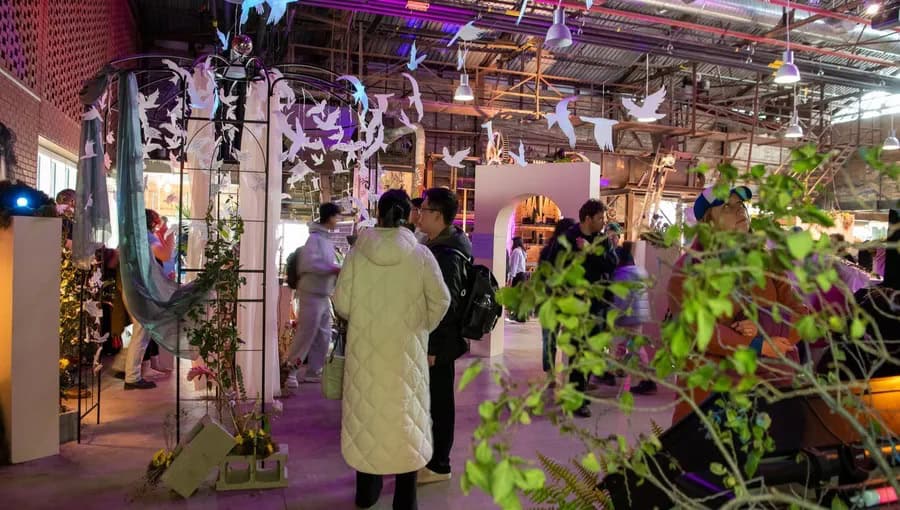 People attending The Good Mourning Festival 2026 in an indoor venue decorated with hanging paper birds and lush greenery, engaging in conversations and exploring exhibits.