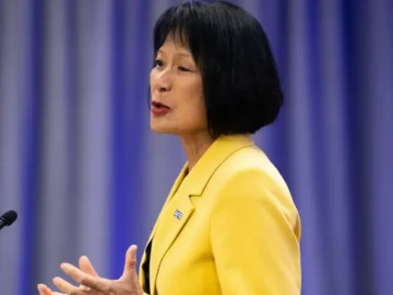 Olivia Chow speaking at a podium in a bright yellow blazer during a press event in Toronto, addressing the audience with a focused expression.
